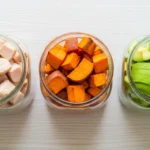 Uma foto de alta qualidade, de cima, mostrando três potes com alimentos representando os macronutrientes: um com peito de frango (proteína), outro com batata-doce (carboidrato) e o terceiro com abacate (gordura). A foto é limpa e organizada. Palavra-chave na descrição: o que são macronutrientes